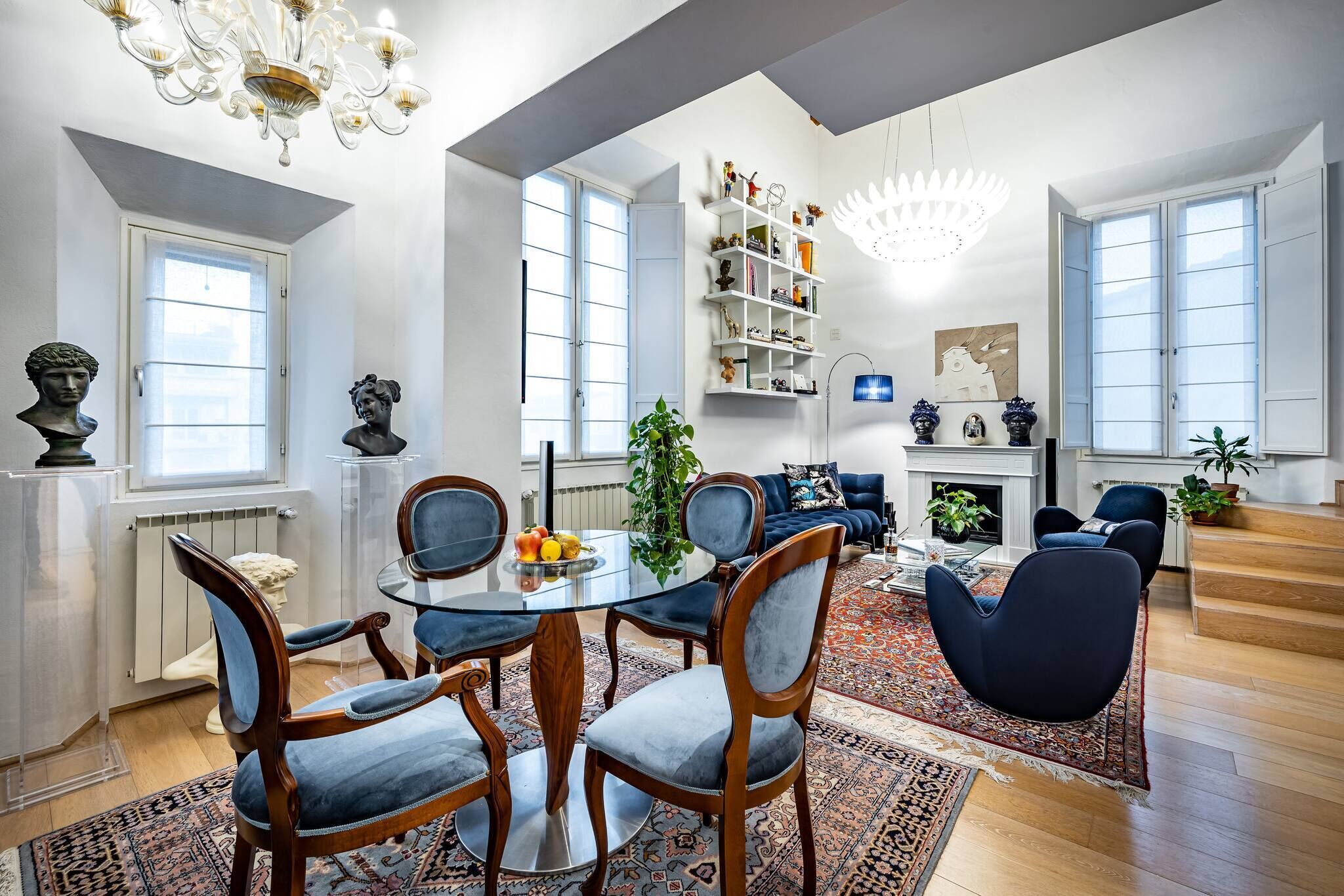 A spacious living room with a round glass table, blue upholstered chairs, a fireplace, and a large bookshelf.