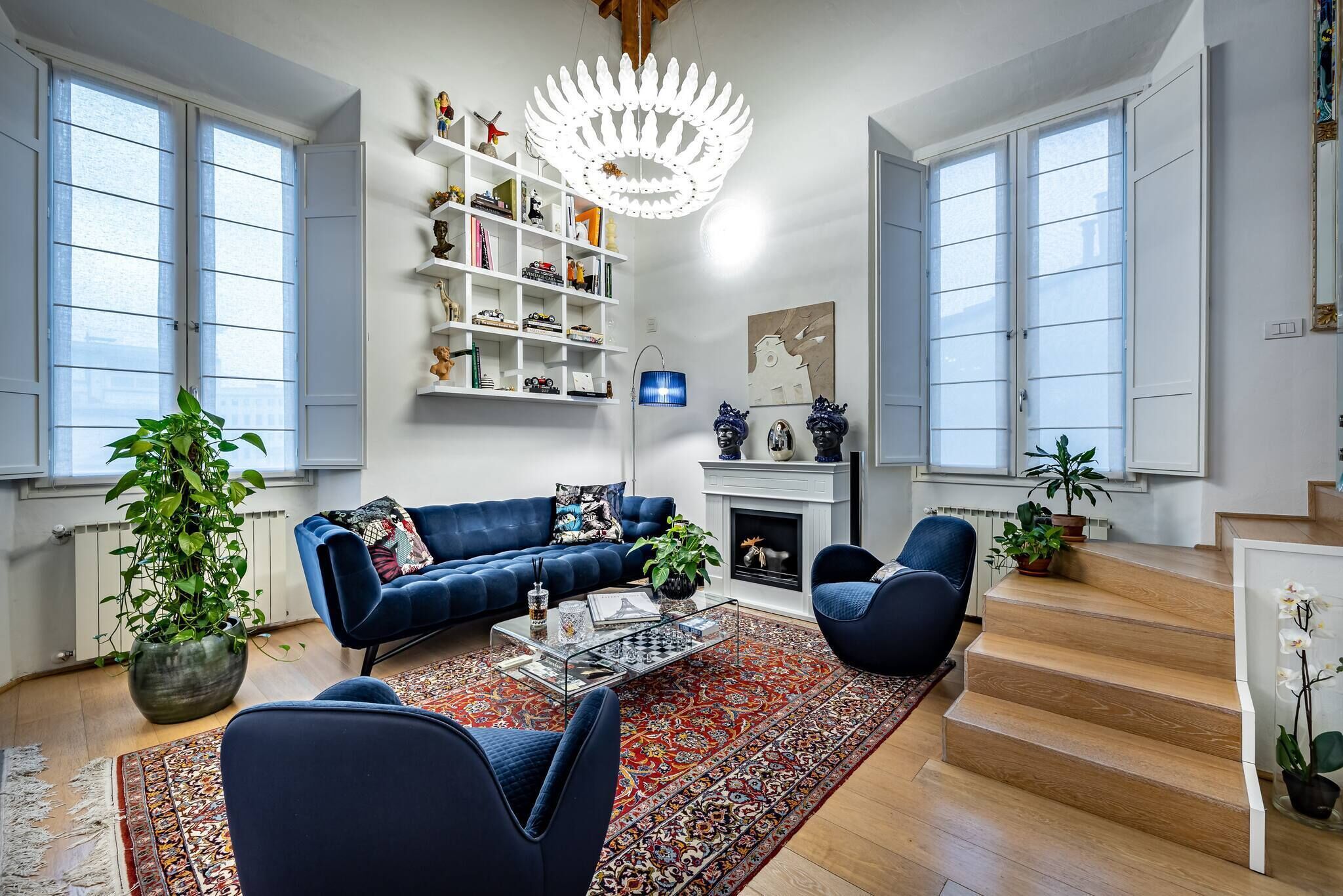 A living room with a blue sofa, a coffee table, and a patterned rug. There are two chairs, a bookshelf, and a fireplace.