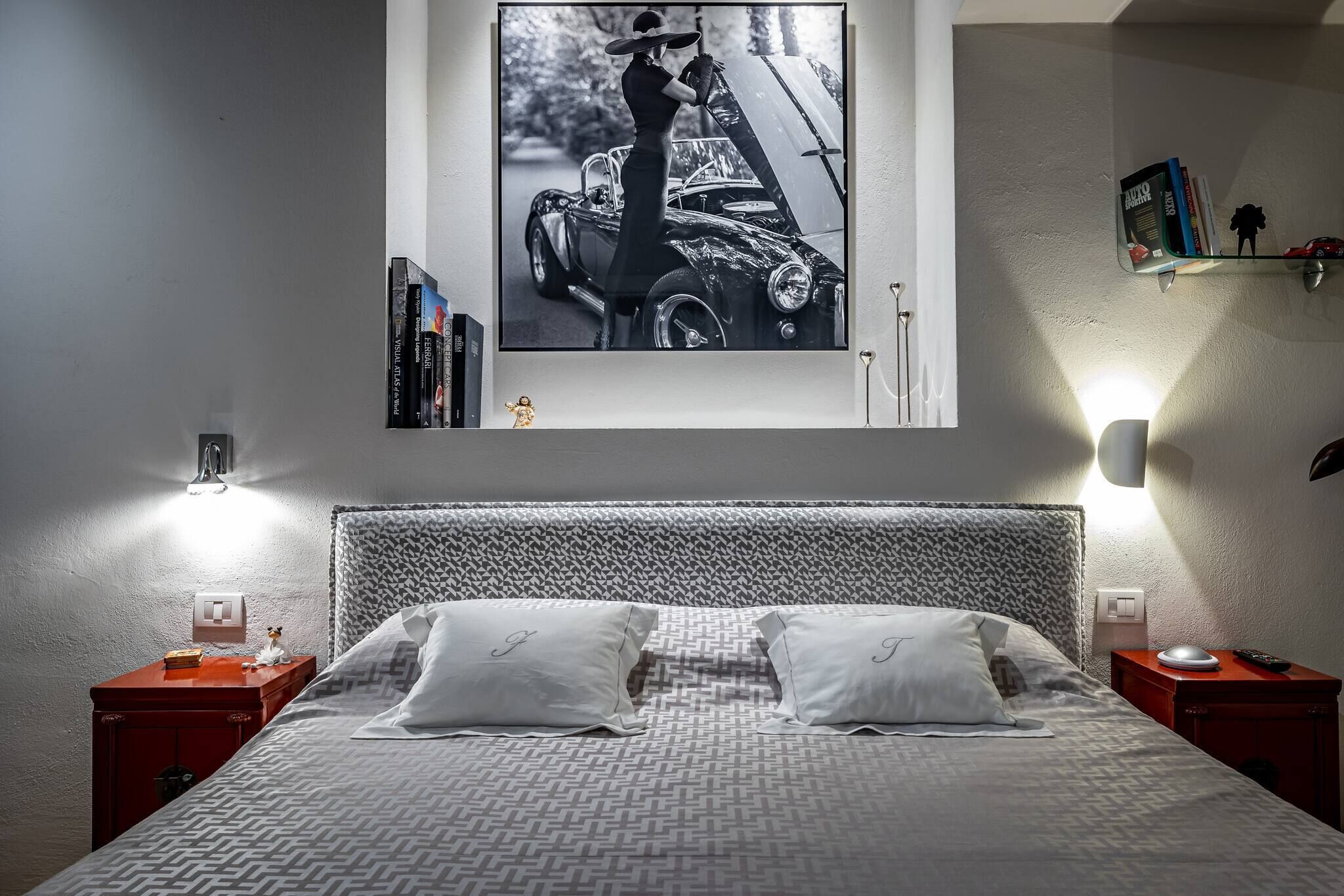 A neatly made bed with a patterned headboard, two bedside tables with lamps, a framed picture of a vintage car on the wall, and a small shelf with books and a figurine.
