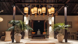 A room with a central seating area, large potted plants, hanging light fixtures, and a wooden ceiling structure.