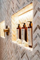A bathroom with a herringbone tile wall, a gold door handle, and three bottles of hair care products on a marble ledge.