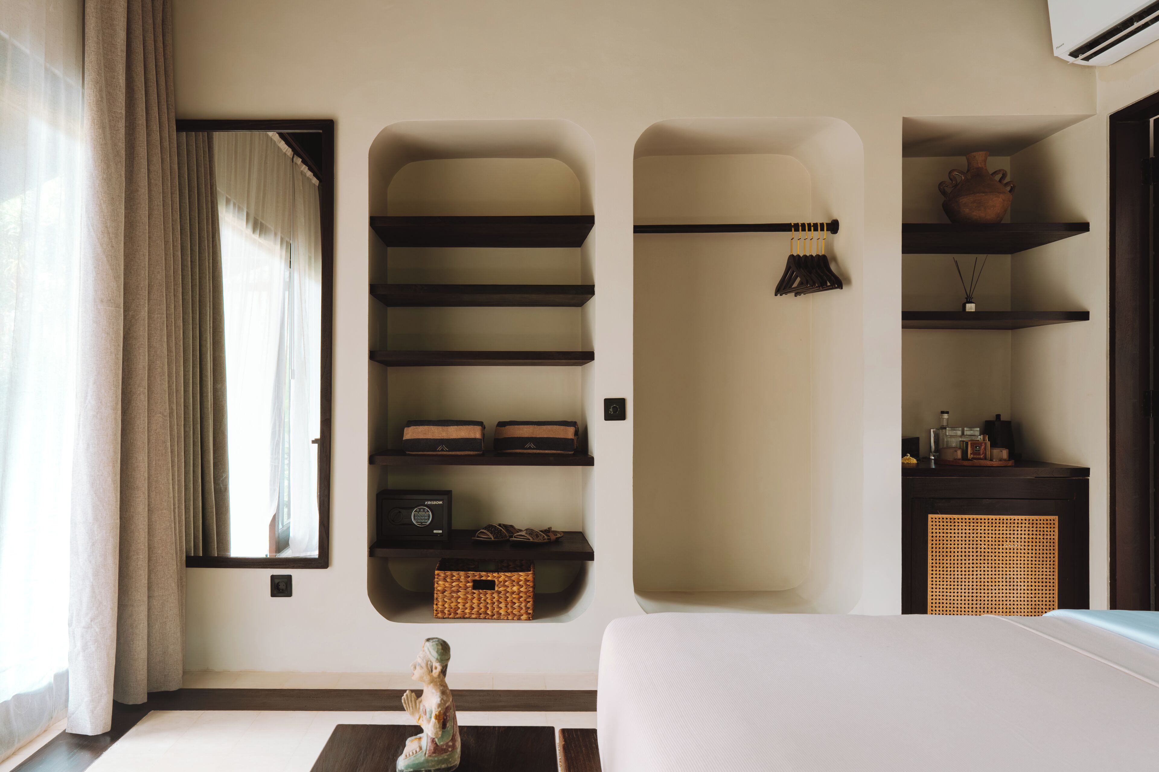 Una habitación con una cama, una ventana con cortinas, estantes empotrados, una cesta de mimbre y una pequeña estatua.