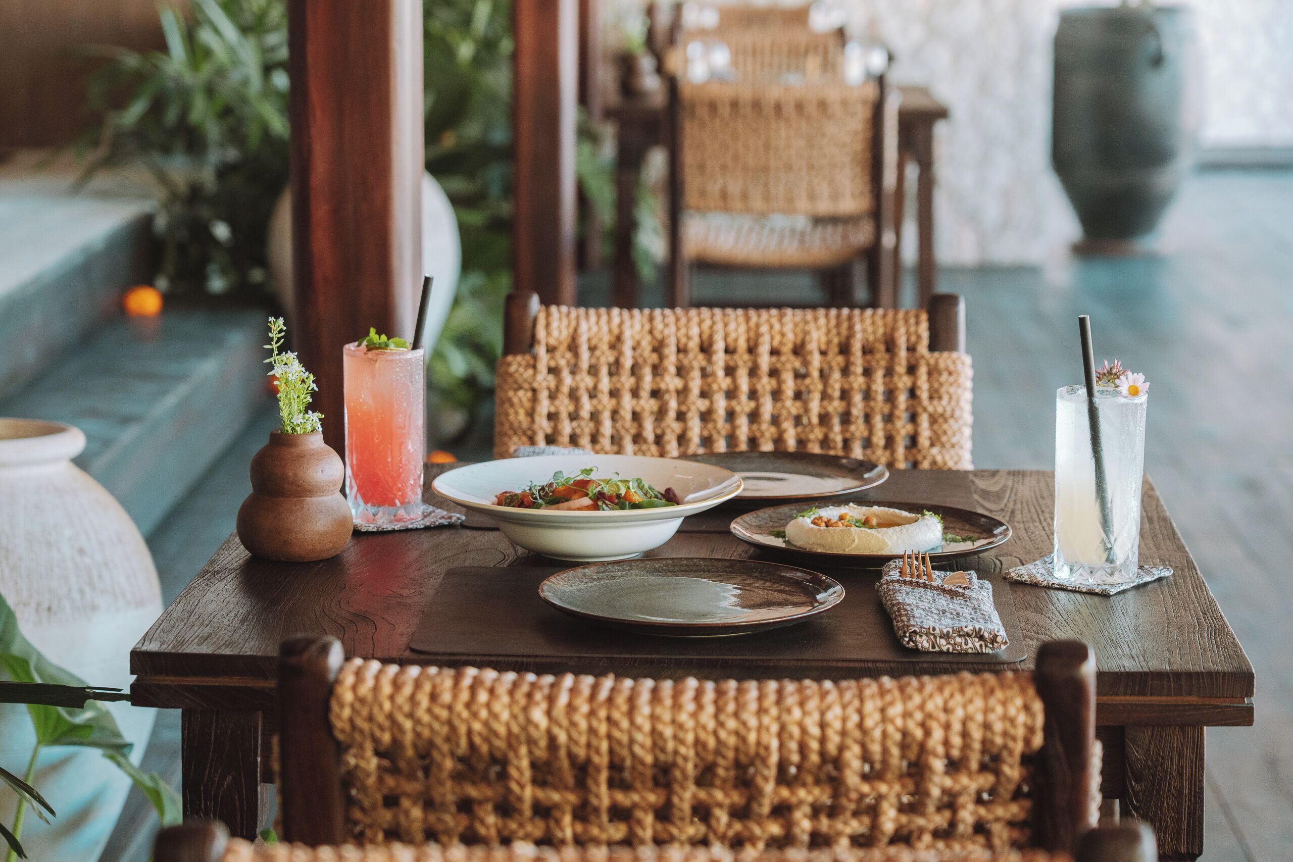 Una mesa de madera con sillas de mimbre, un tazón de ensalada y dos bebidas con pajitas.
