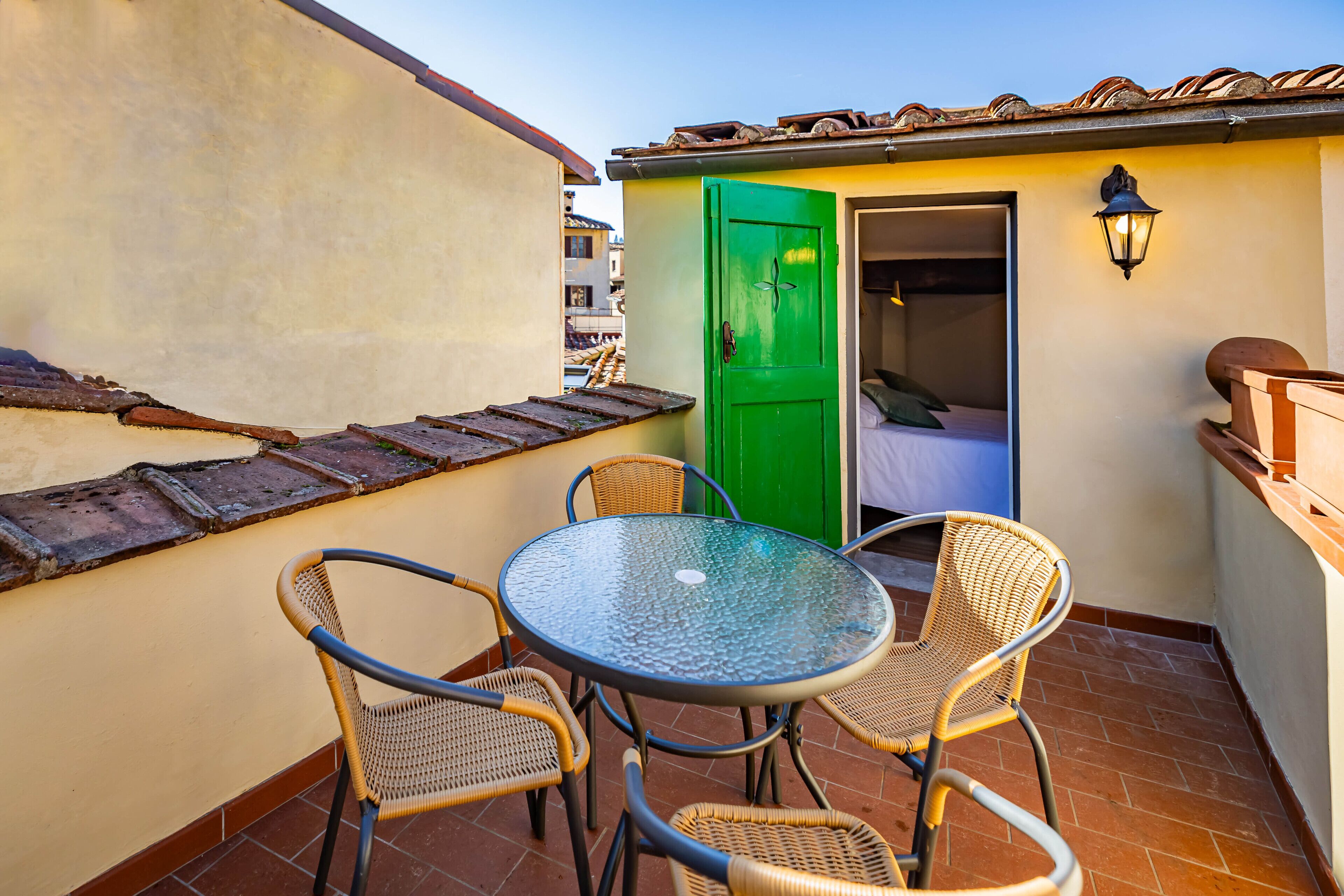 A small balcony with a round glass table and wicker chairs, a green door, and a tiled roof.