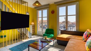 Ein hellgelbes Zimmer mit einem großen Fenster, einem grünen Sessel, einer Couch, einem Flachbildfernseher und einer Treppe.