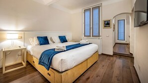 A bedroom with a wooden bed, white bedding, a blue throw, a nightstand with a lamp, and a window with a view. - Cantoria (Florence)