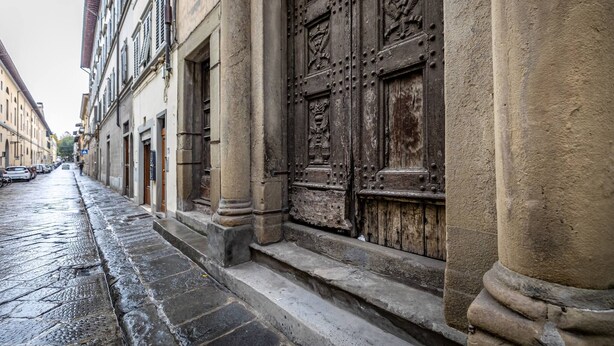 A narrow street with historic buildings, a large ornate wooden door, and a wet cobblestone path. - Cantoria (Florence)