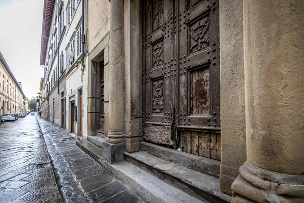A narrow street with historic buildings, a large ornate wooden door, and a wet cobblestone path. - Cantoria (Florence)