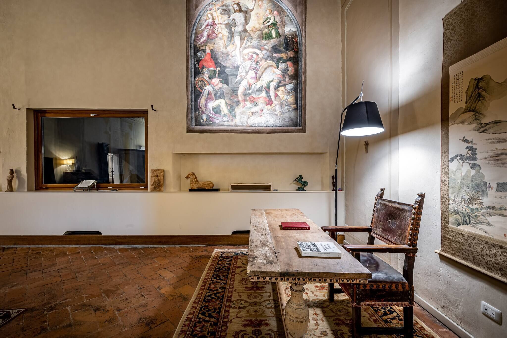 A room with a large stained glass window, a wooden table with chairs, a patterned rug, and a traditional Chinese painting on the wall.