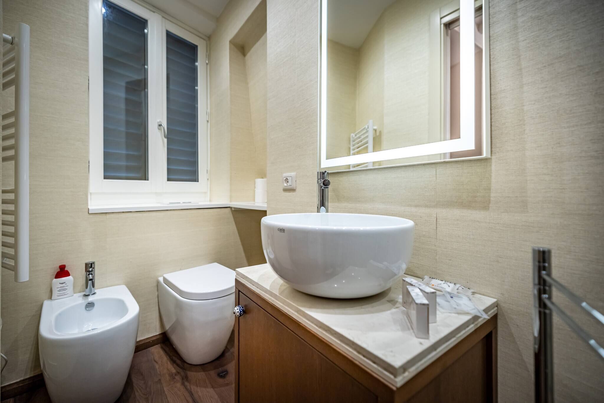 A bathroom with a white sink, toilet, and bidet, wooden vanity, and a window with blinds.