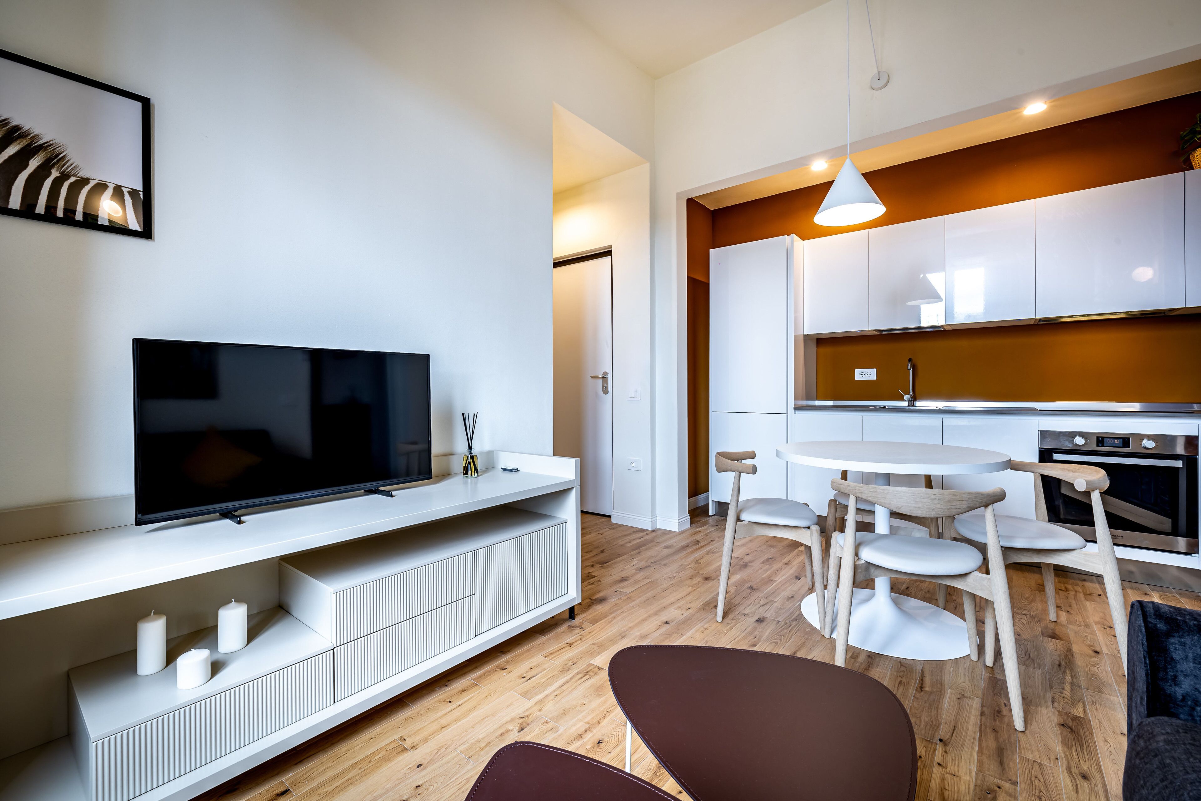 Una sala moderna con una televisión de pantalla plana, mueble blanco de entretenimiento, piso de madera y una zona de comedor con una mesa redonda y sillas.