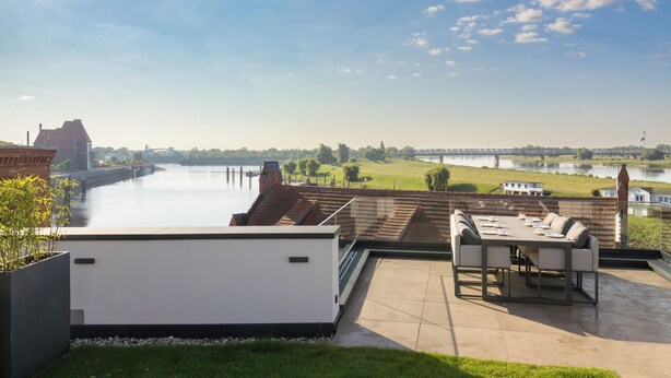 A rooftop terrace with a view of a river, a dining table with chairs, and a modern building in the background. - ELBE.6 – Apartment II | 5-star right on the Elbe (Wittenberge)