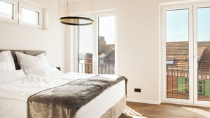 A bedroom with a large bed, a hanging light fixture, and a view of buildings through a sliding door. - ELBE.6 – Apartment II | 5-star right on the Elbe (Wittenberge)