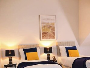 Hotellihuone, jossa on kaksi sänkyä. Jokaisessa sängyssä on pääty, yöpöydällä on lamppu ja seinällä on taulu.