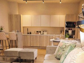 Eine moderne Küche mit hellen Holzschränken, ein Essbereich mit Tisch und Stühlen sowie ein Wohnbereich mit Sofa und Hocker.