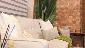 Een witte bank met gestreepte kussens, een groene plaid en een grote groene plant in een kamer met een bakstenen muur en gestreepte jaloezieën.