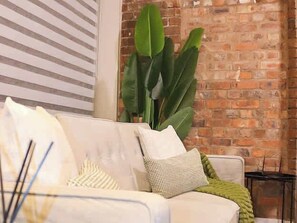 A white sofa with striped pillows, a green throw blanket, and a large green plant in a room with a brick wall and striped blinds. - Effortless Stays at Luxury Birmingham 2BR Sleeps 6 (Deritend)