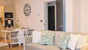 Een woonkamer met een bank, een glazen salontafel, een eettafel met stoelen en een klok aan de muur.