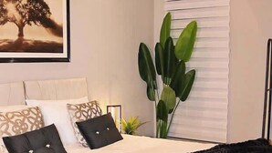 Een slaapkamer met een bed, kussens, een plant en een raam met jaloezieën.