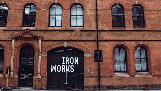 Een gebouw van rode baksteen met gebogen ramen en een grote zwarte deur waarop 'IRON WORKS' staat.