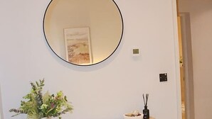 Een kamer met een ronde spiegel aan de muur, een lijst met afbeelding, een potplant en een klein tafeltje met een decoratief object.