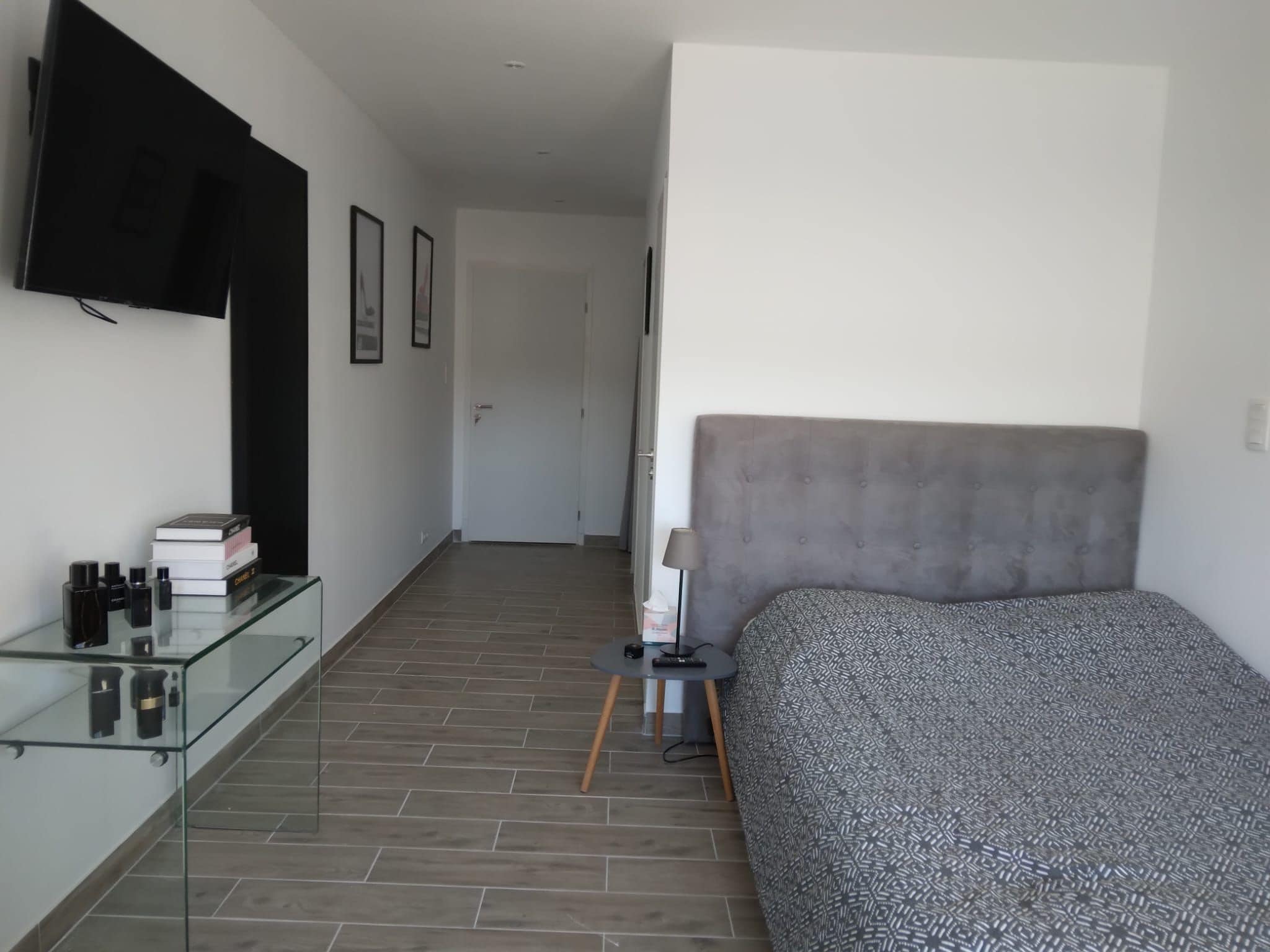 A bedroom with a bed, a TV mounted on the wall, a bedside table with a lamp, and a glass cabinet with toiletries.