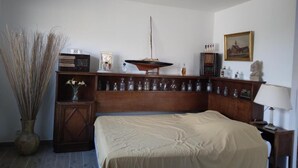 Un dormitorio con una cama, un armario de madera, un florero con plantas secas, una mesita de noche con una lámpara y un cuadro en la pared.