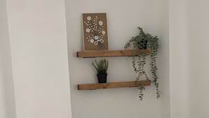 Un lit avec une literie blanche, un coussin turquoise et un coussin blanc. On y voit deux étagères en bois avec des plantes et un tableau encadré.