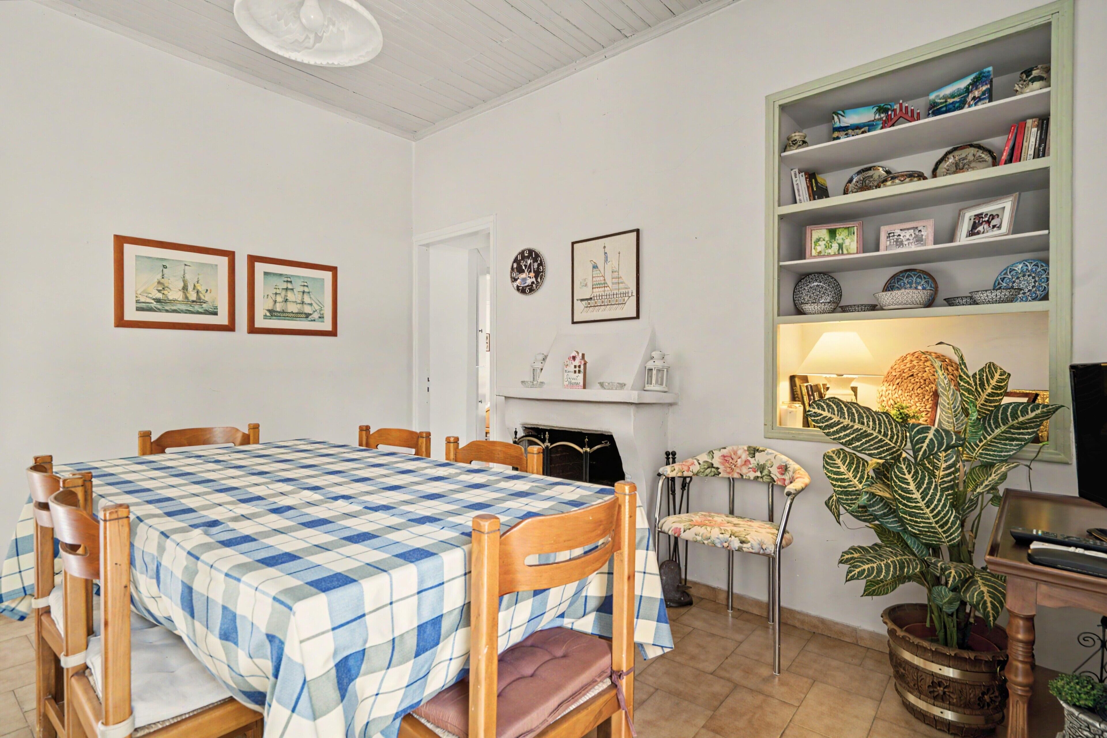 Un coin repas avec une nappe à carreaux, des chaises en bois, une cheminée et une bibliothèque agrémentée d’objets décoratifs.