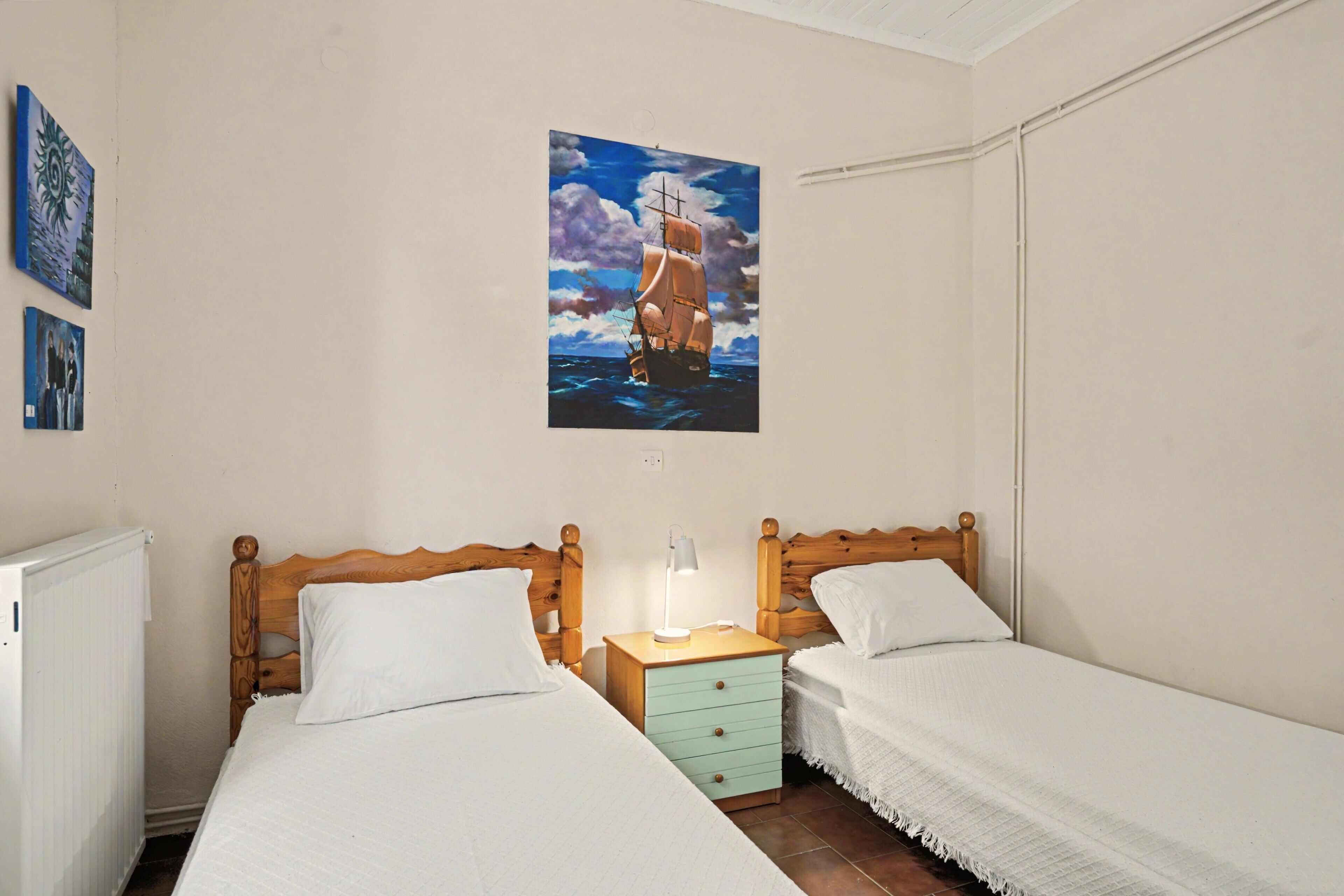 A bedroom with two beds, a nightstand with a lamp, and a painting of a ship on the wall.