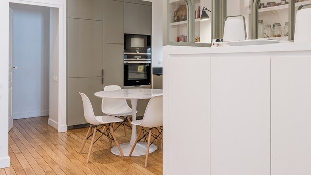 Une cuisine moderne avec des meubles blancs, une table à manger avec des chaises et une grande fenêtre donnant sur l’intérieur.