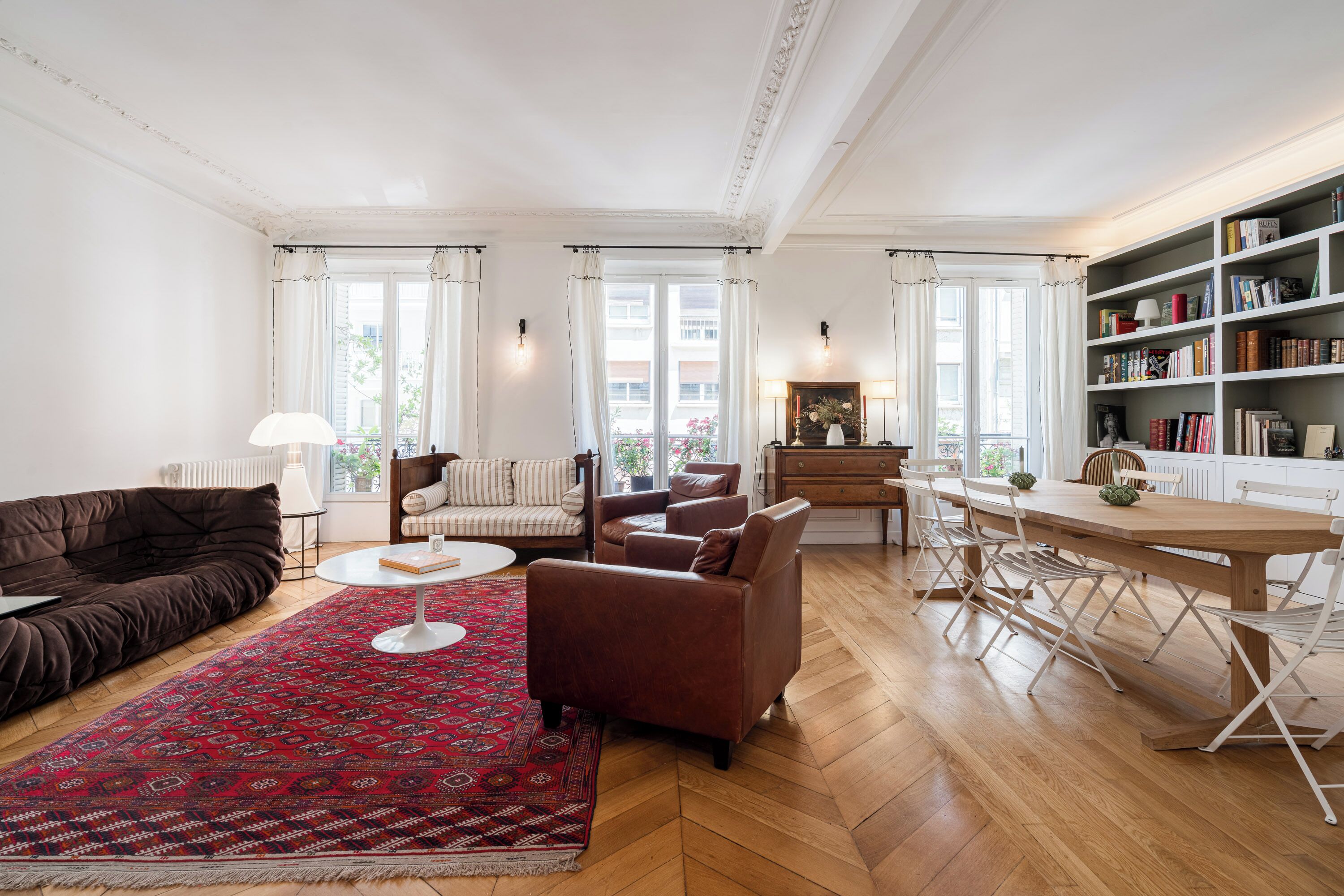 A spacious living room with a large window, a fireplace, a bookshelf, and a dining area.