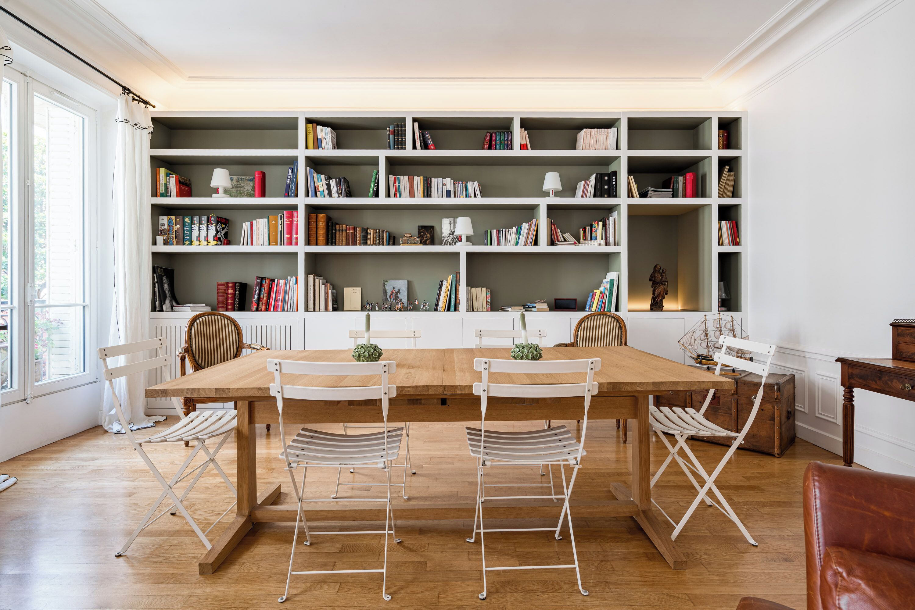 A dining area with a wooden table and white chairs, a bookshelf with books and decorative items, and a window with curtains.