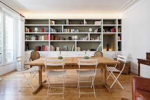 A dining area with a wooden table and white chairs, a bookshelf with books and decorative items, and a window with curtains. - Avenue Foch Retreat, by Veeve (Paris)