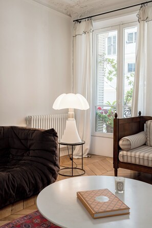 Ein gemütliches Zimmer mit einem dunkelbraunen Sofa, einem runden weißen Tisch mit Buch und Kerze, einer Stehlampe und einem Fenster mit Vorhängen.