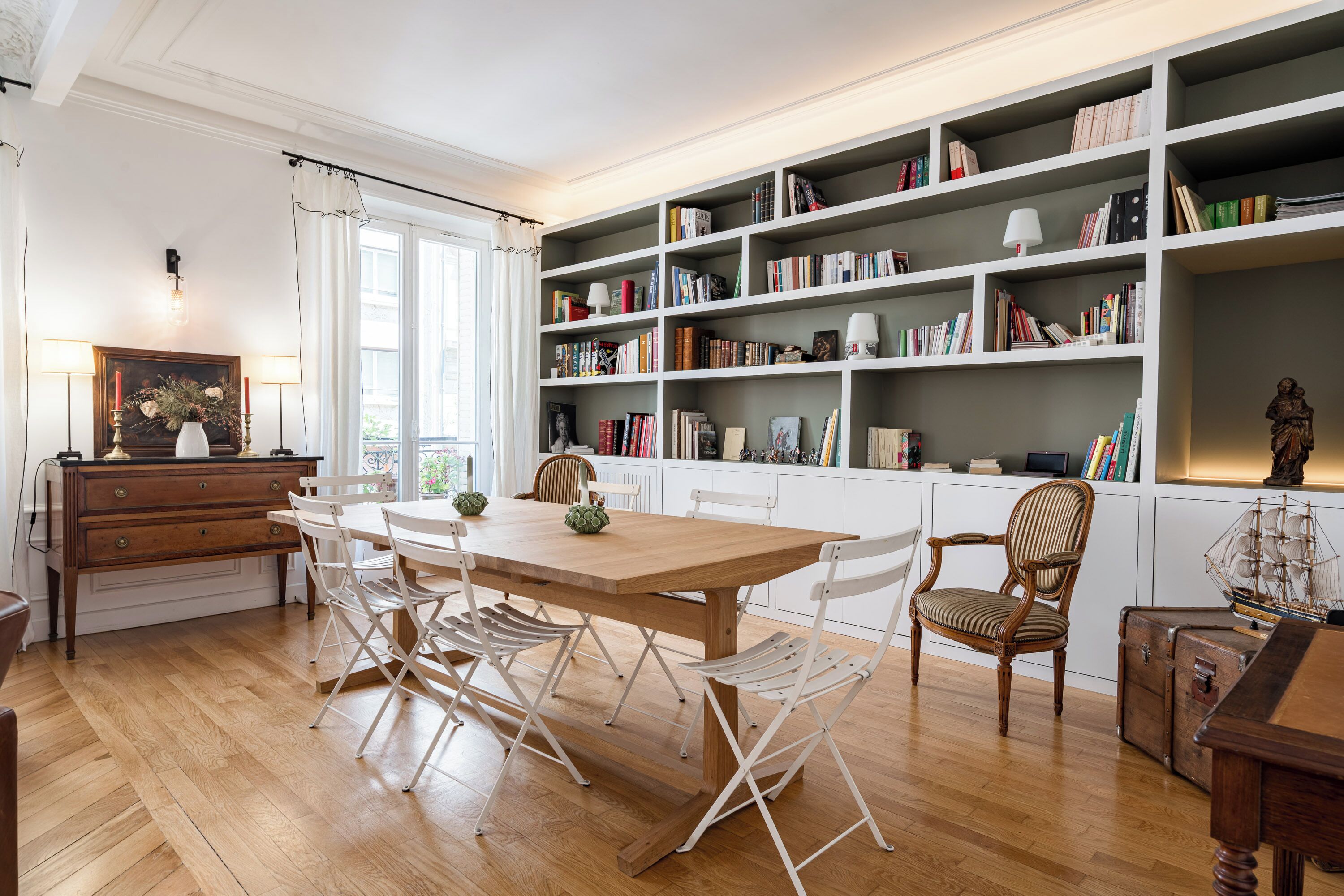 A dining area with a wooden table, white chairs, a bookshelf filled with books, and a decorative vase.