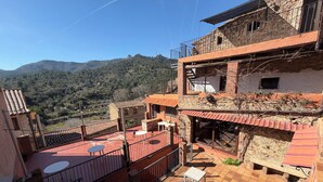 A terrace with outdoor seating, a stone building with a balcony, and a mountainous backdrop.