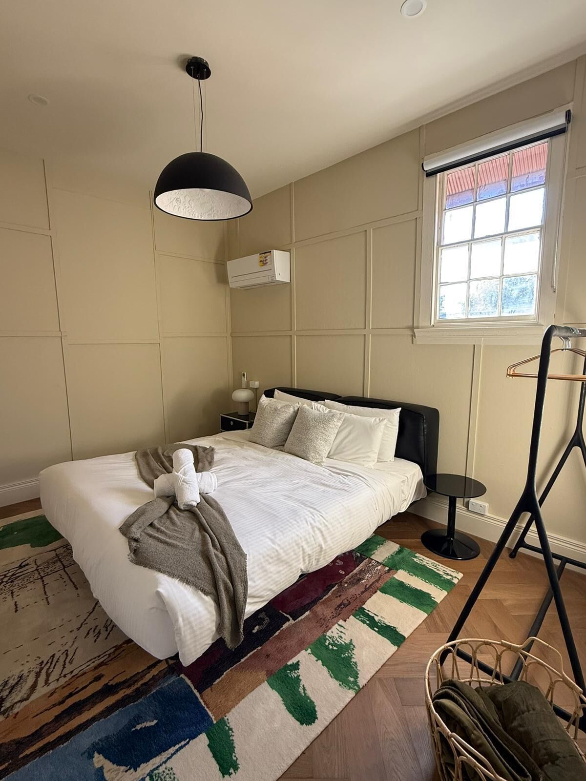 Une chambre à coucher bien rangée, avec un lit, des tables de chevet, un luminaire suspendu, une fenêtre avec des stores et un ventilateur sur pied.