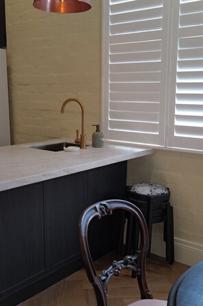 A kitchen with a copper faucet, a sink, a window with blinds, a wooden chair, and a stool. - The Fable Fitzroy Melbourne (With Parking) (Fitzroy)