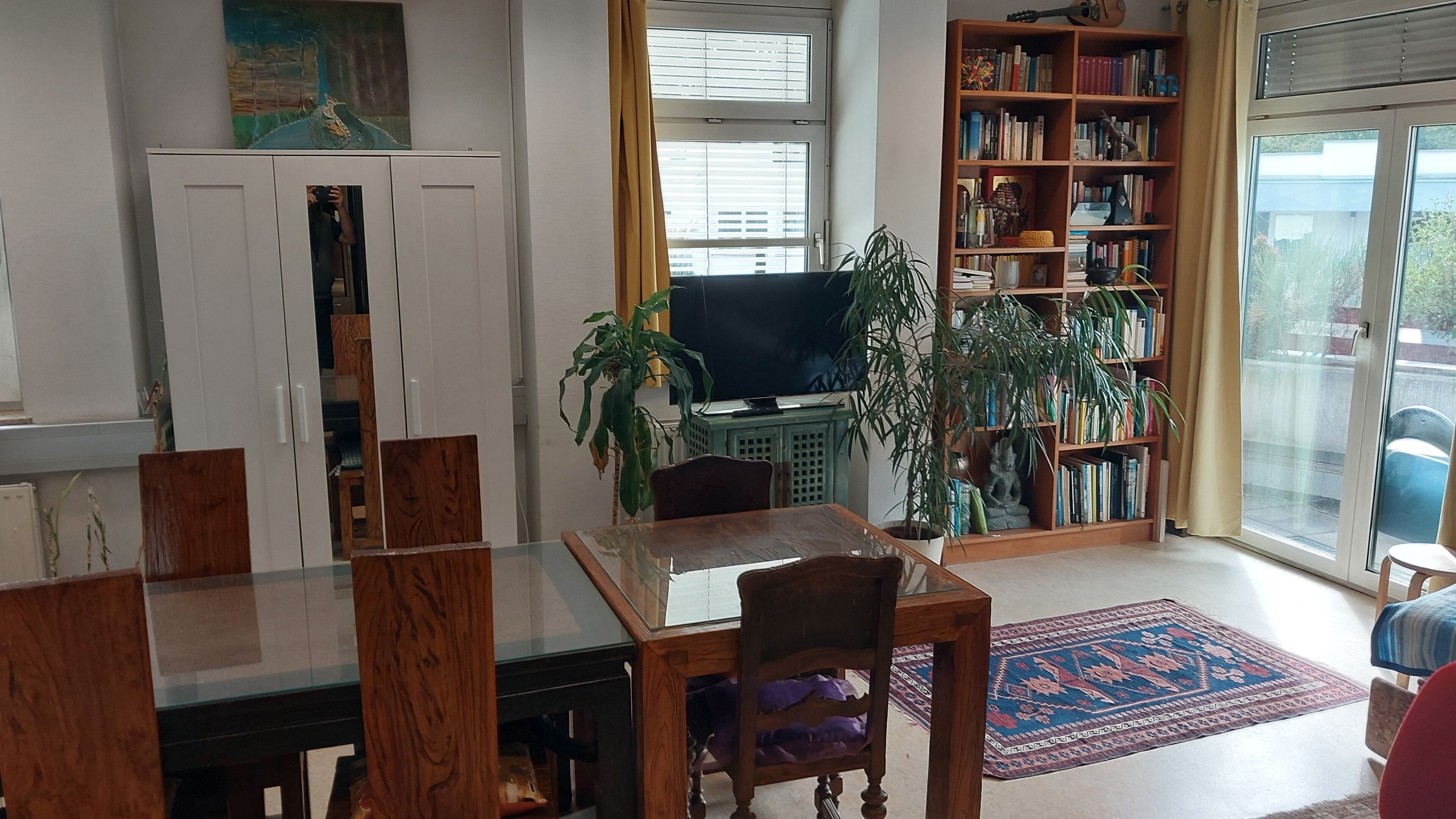 Una sala de estar con una mesa de comedor, sillas, una estantería, un televisor y un amplio ventanal.