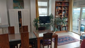 Ein Wohnzimmer mit Esstisch, Stühlen, einem Bücherregal, einem Fernseher und einem großen Fenster.