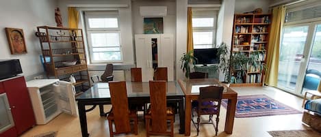 A dining area with a glass table and wooden chairs, a bookshelf, a television, and a large window with curtains.