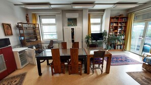 Ein Essbereich mit einem Glastisch, Holzstühlen, einem Bücherregal, einem Fernseher und einem großen Fenster mit Vorhängen.