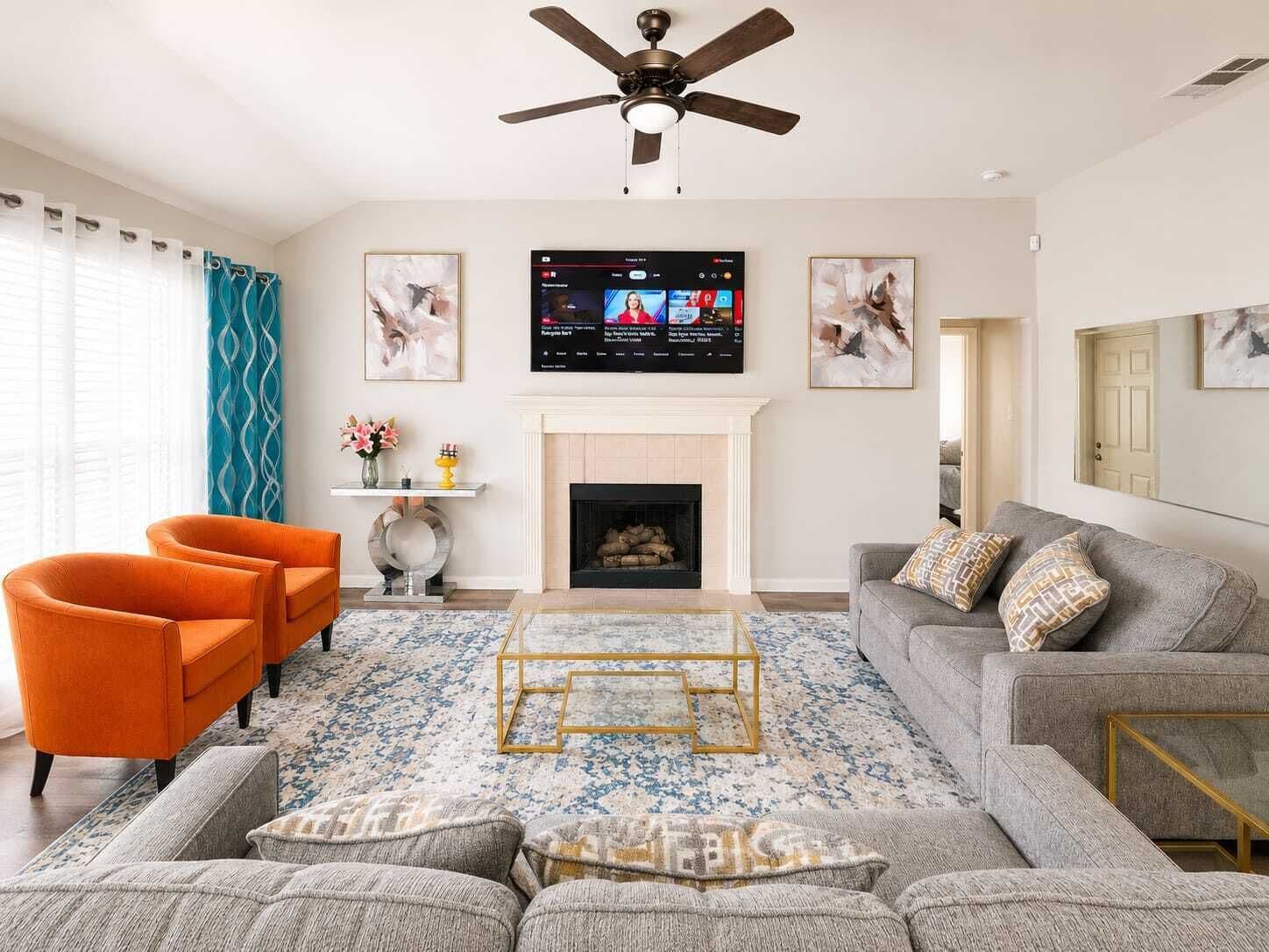 A living room with a fireplace, a ceiling fan, a flat-screen TV, and a large window with curtains.
