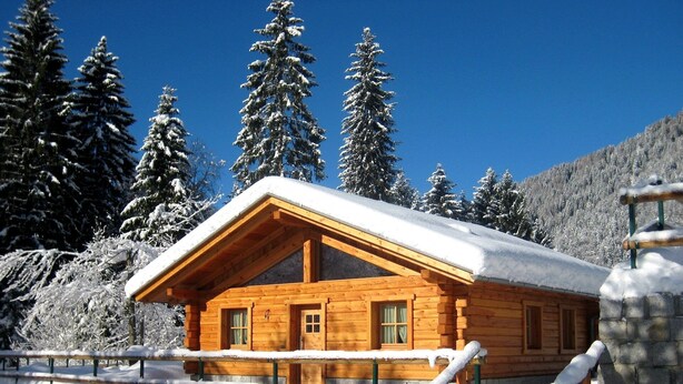 Een houten hut met een sneeuwbedekte dak, omgeven door sneeuwzware bomen en een bergachtig landschap.