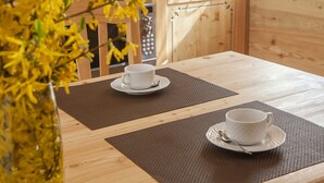 Een houten eettafel met twee witte kopjes en schotels op placemats, een keuken met houten kasten, en een uitzicht op gele bloemen buiten.