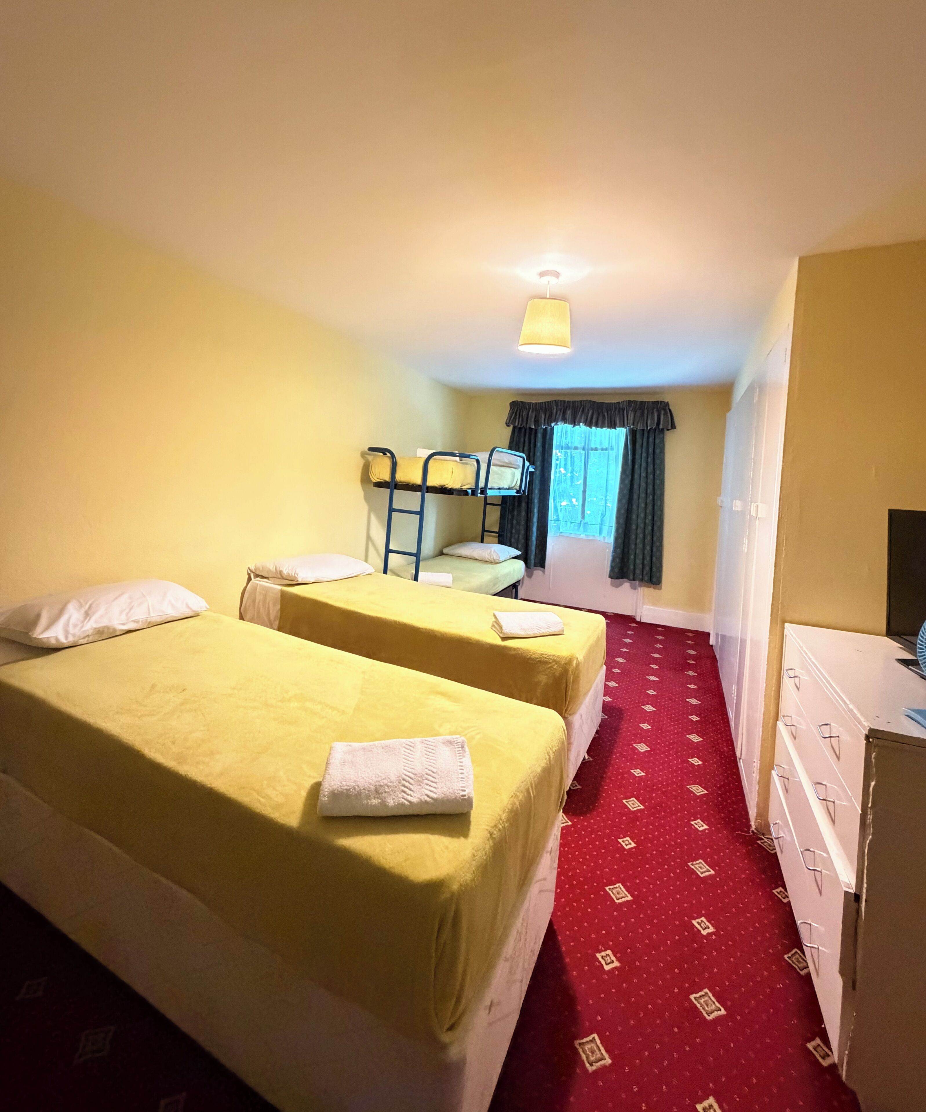 Habitación de hotel con dos camas, un escritorio, una silla, una ventana con cortinas y una alfombra roja.
