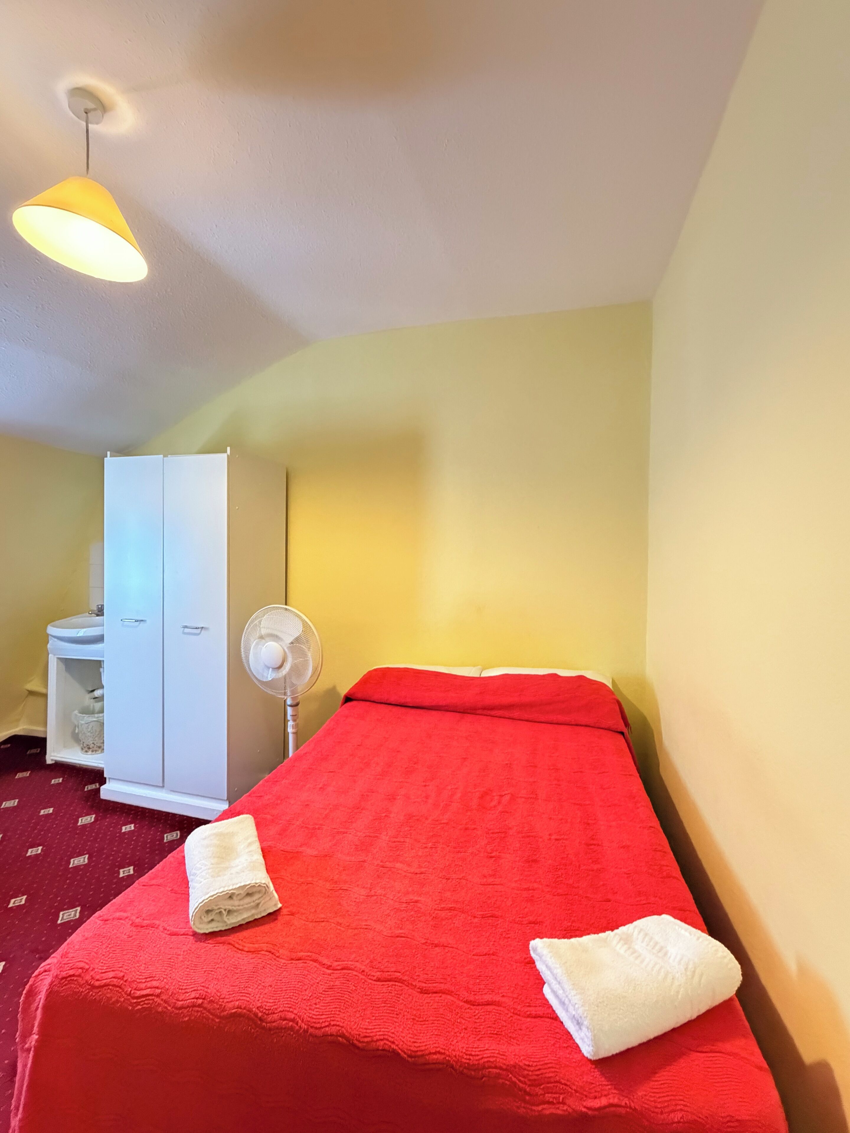 Un lit simple avec une couverture rouge, un ventilateur blanc et une armoire dans une pièce aux murs jaunes.