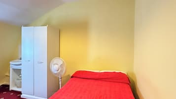 Un lit simple avec une couverture rouge, un ventilateur blanc et une armoire dans une pièce aux murs jaunes.
