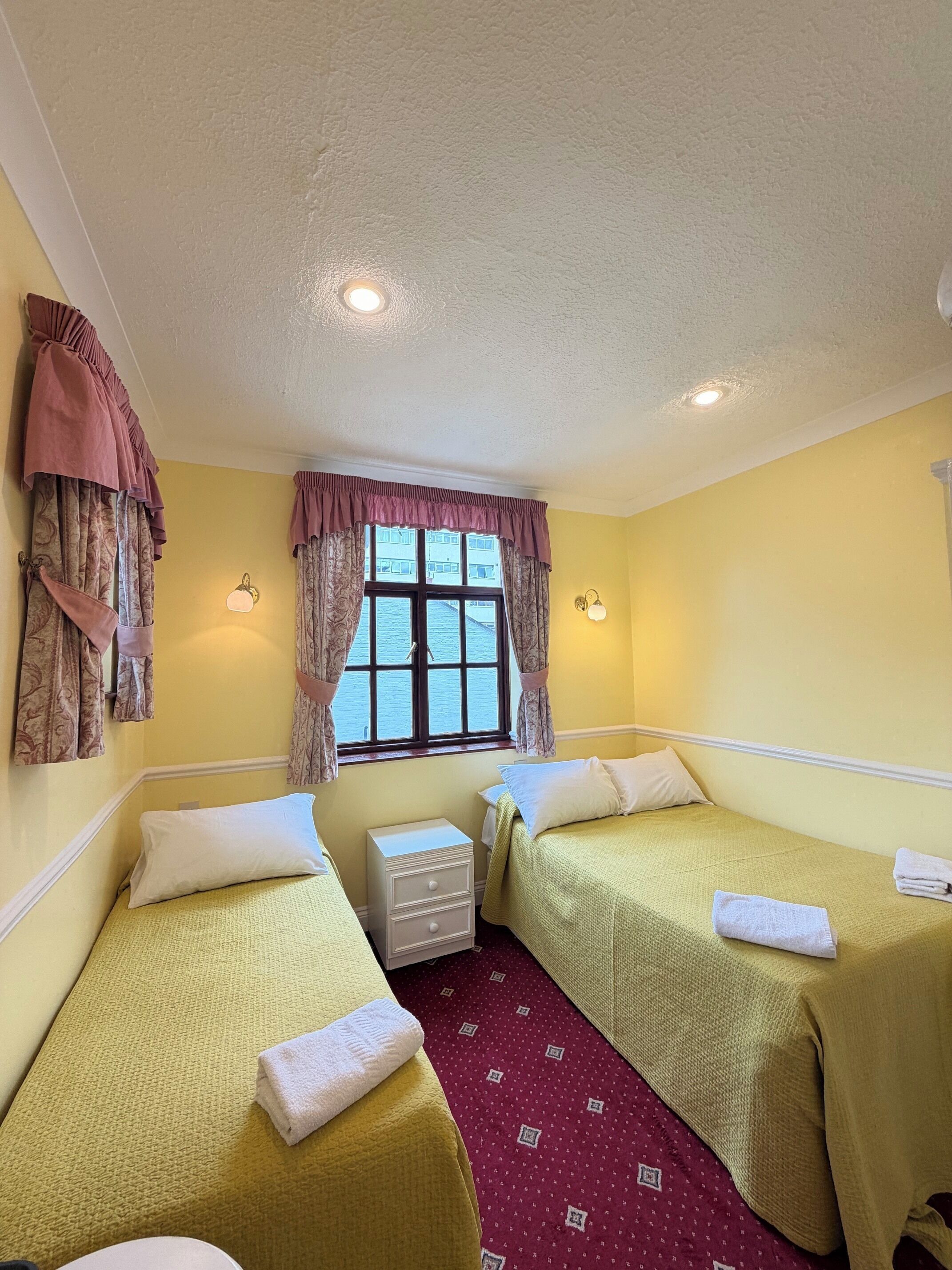A room with two single beds, a window with curtains, a small bedside table, and a wall-mounted lamp.